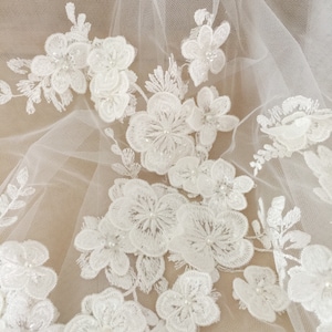 May include: White floral lace fabric with pearl embellishments. The lace is delicate and intricate, with a soft, feminine look.