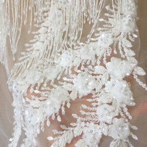 May include: White beaded lace fabric with a floral design and fringe. The fabric is sheer and delicate, with a soft, flowing texture.