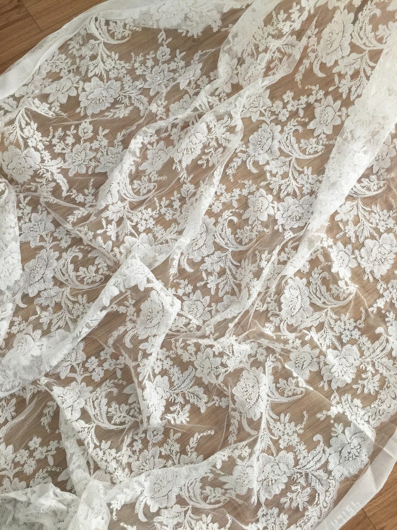 May include: White lace fabric with a delicate floral pattern. The lace is sheer and has a soft, flowing texture.