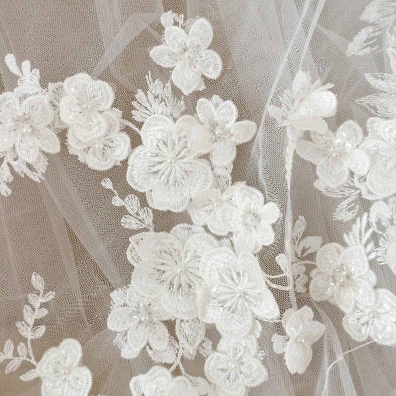 May include: White floral lace fabric with pearl embellishments. The lace is delicate and intricate, with a variety of flower shapes and sizes. The pearls are small and evenly spaced, adding a touch of elegance to the fabric.