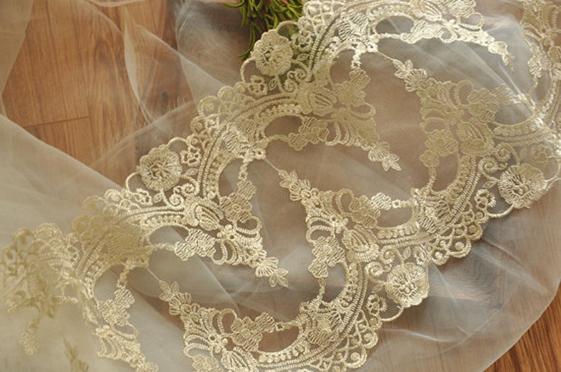 Puede incluir: Borde de encaje dorado con un delicado patr&oacute;n floral sobre una tela blanca transparente.
