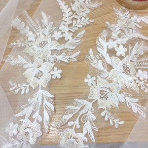 Pode incluir: Close-up de apliques de renda floral branca com bordados detalhados. O design apresenta flores e folhas delicadas, com um tom branco-sujo suave. A renda é colocada sobre um tecido branco transparente, criando um efeito em camadas. Uma almofada de alfinetes vermelha e branca está no fundo.