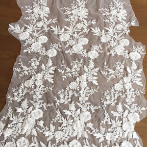 May include: White floral lace fabric with a delicate, intricate design. The lace features raised, three-dimensional flowers and leaves on a sheer mesh background. The fabric has a scalloped edge.