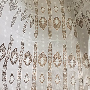 Puede incluir: Tela de encaje de algod&oacute;n blanco con un patr&oacute;n delicado e intrincado. El encaje presenta un dise&ntilde;o repetitivo de l&iacute;neas verticales con motivos florales.