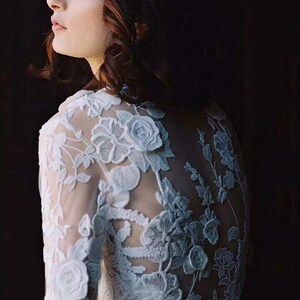 May include: A white lace wedding dress with long sleeves and a floral design. The dress has a sheer back with a floral pattern.