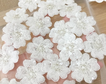 May include: White flower-shaped fabric embellishments with sequins and beads. The embellishments are on a sheer white fabric background.