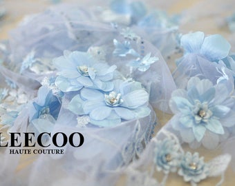 Puede incluir: Tela de encaje floral azul claro con una textura delicada y transparente. La tela presenta flores en 3D y un brillo sutil. La tela está etiquetada con "LEECOO HAUTE COUTURE".