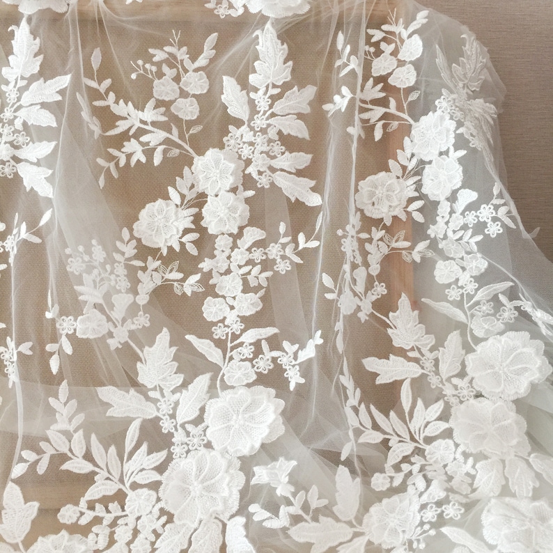 May include: White floral lace fabric with a delicate pattern of flowers and leaves. The lace is on a sheer, white mesh background.