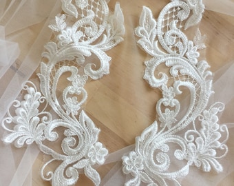 May include: Two white lace appliques with intricate floral designs. The appliques are delicate and would be perfect for adding a touch of elegance to a wedding dress or other garment.