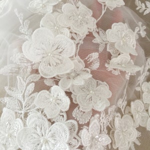 May include: White floral lace fabric with pearl embellishments. The lace is delicate and intricate, with a floral pattern. The fabric is perfect for adding a touch of elegance to any project.