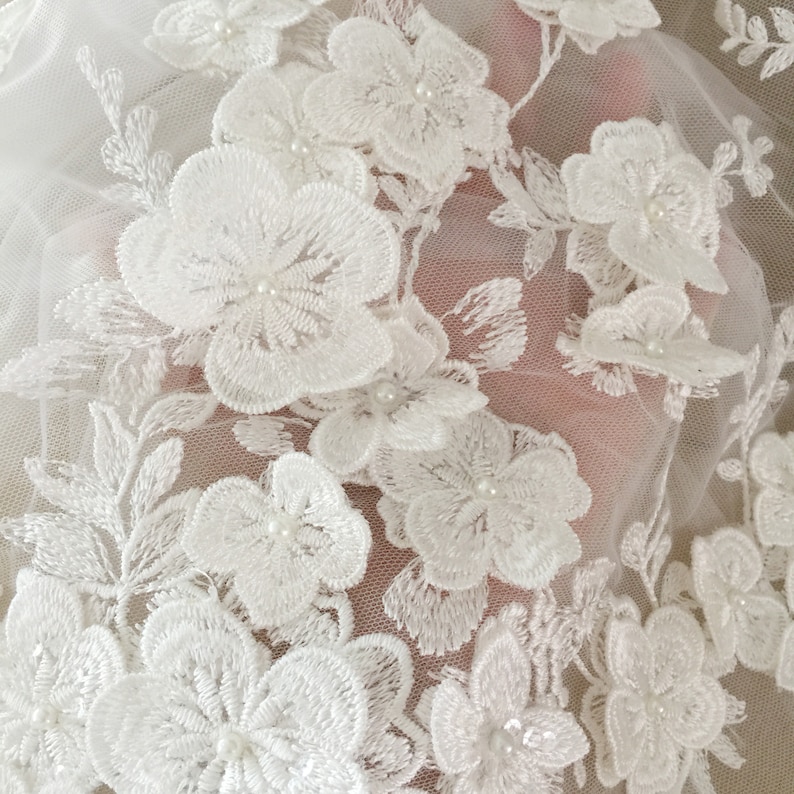 May include: White floral lace fabric with pearl embellishments. The lace is delicate and intricate, with a floral pattern. The fabric is perfect for adding a touch of elegance to any project.