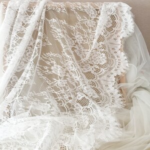 May include: White lace fabric with a delicate floral pattern. The lace has a scalloped edge and is soft and sheer.