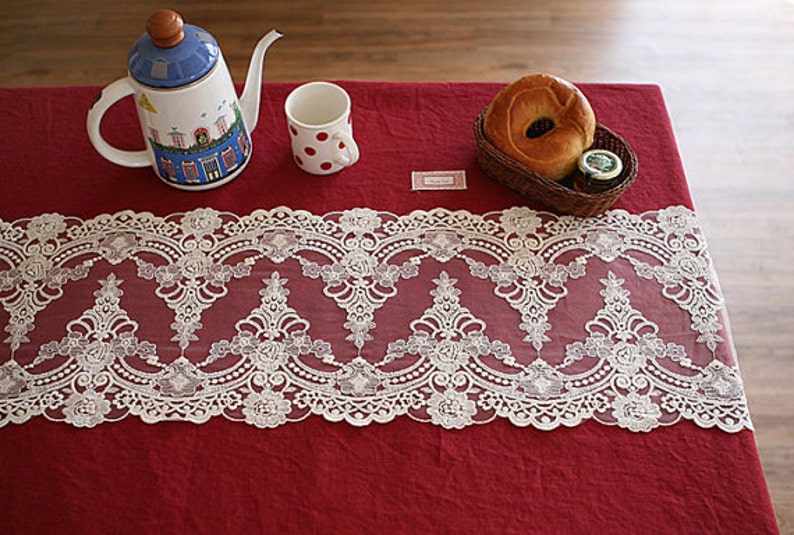 Puede incluir: Borde de encaje blanco con un delicado patr&oacute;n floral, colocado sobre un mantel rojo con una tetera, una taza y un pastel.