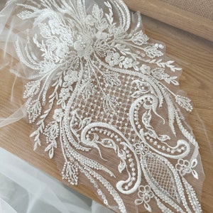May include: An intricate white lace applique with floral and swirling patterns. The design features beaded accents, sequins, and delicate embroidery on a sheer mesh backing. This decorative element is suitable for bridal or formal wear.