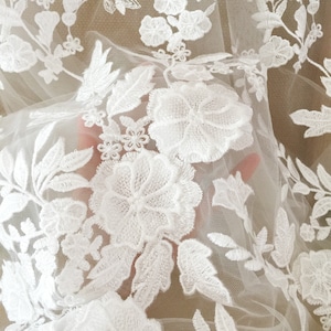 May include: White floral lace fabric with a delicate pattern of flowers and leaves. The lace is sheer and has a soft, romantic feel.