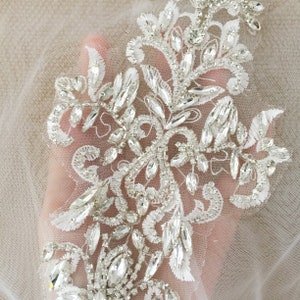 May include: A delicate white lace applique with a floral design embellished with sparkling clear rhinestones. The applique is perfect for adding a touch of elegance to wedding dresses, bridal accessories, or other special occasion garments.