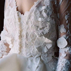 May include: A white lace wedding dress with a high neckline and long sleeves. The dress is adorned with delicate floral embroidery and 3D floral appliques.