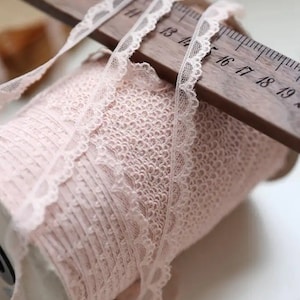 May include: A spool of pale pink lace trim with a scalloped edge. The lace is wound around a white spool and is being measured with a wooden ruler marked in inches.