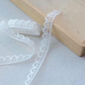 May include: Delicate white lace trim with a scalloped edge and a dotted pattern. The lace is rolled up and laid out on a light-colored surface, showcasing its intricate design. Ideal for sewing and craft projects.