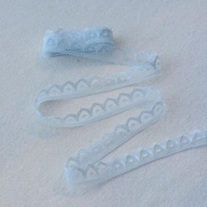 May include: A roll of light blue lace ribbon with a scalloped edge. The ribbon is unrolled, showing the delicate, patterned design. The lace is a soft, pastel blue color.