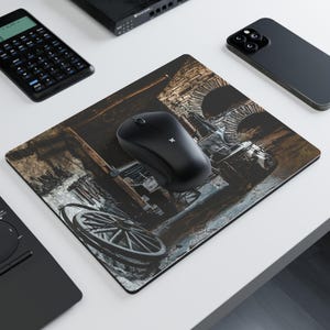 Peut inclure: Une souris d'ordinateur sans fil noire est posée sur un tapis de souris rectangulaire. Le tapis de souris présente une image détaillée, sépia, d'une vieille roue de chariot et d'une structure en pierre. D'autres articles technologiques sont également visibles.