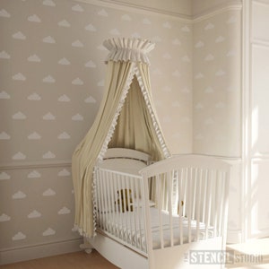 May include: A white cot with a canopy draped over it. The canopy is made of a cream-coloured fabric with white lace trim. The wall behind the cot is painted a light beige colour and features a white cloud pattern.