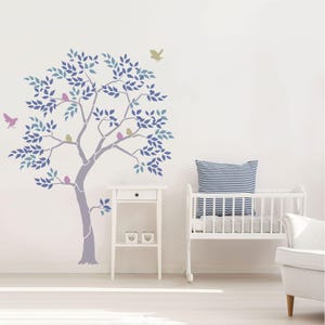 May include: A wall decal featuring a stylized tree with blue and purple leaves, and birds perched on its branches. The tree is painted on a white wall, with a small white side table and a crib in the background. The overall theme is nature and nursery decor.
