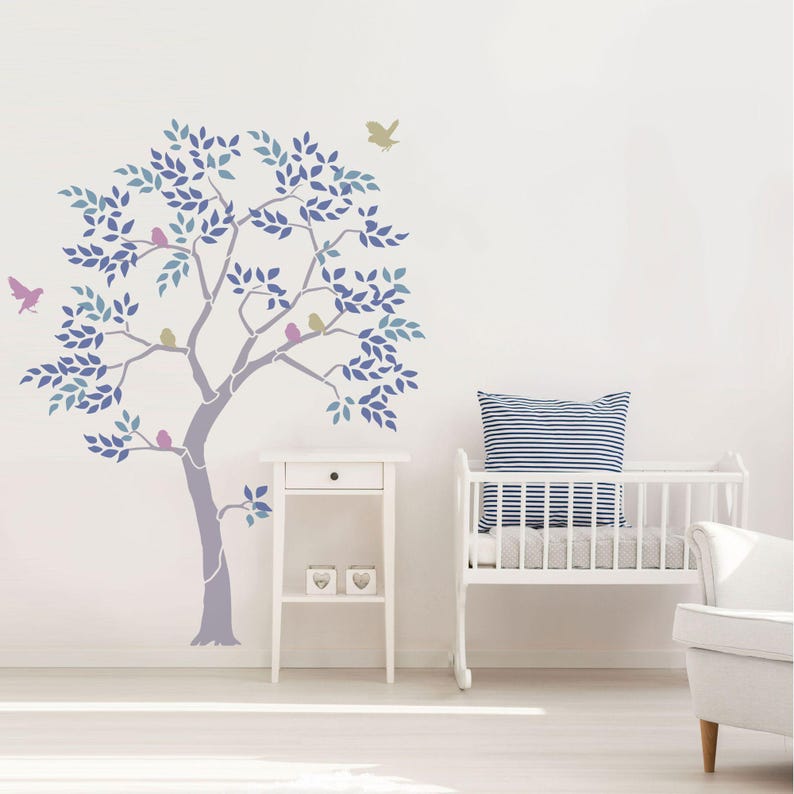 May include: A wall decal featuring a stylized tree with blue and purple leaves, and birds perched on its branches. The tree is painted on a white wall, with a small white side table and a crib in the background. The overall theme is nature and nursery decor.