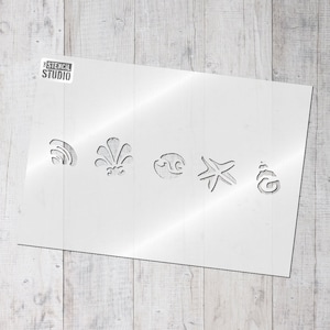 May include: A white stencil sheet with five cut-out designs: a wave, a seashell, a swirl, a starfish, and a seashell. The stencil sheet is on a light wood background.