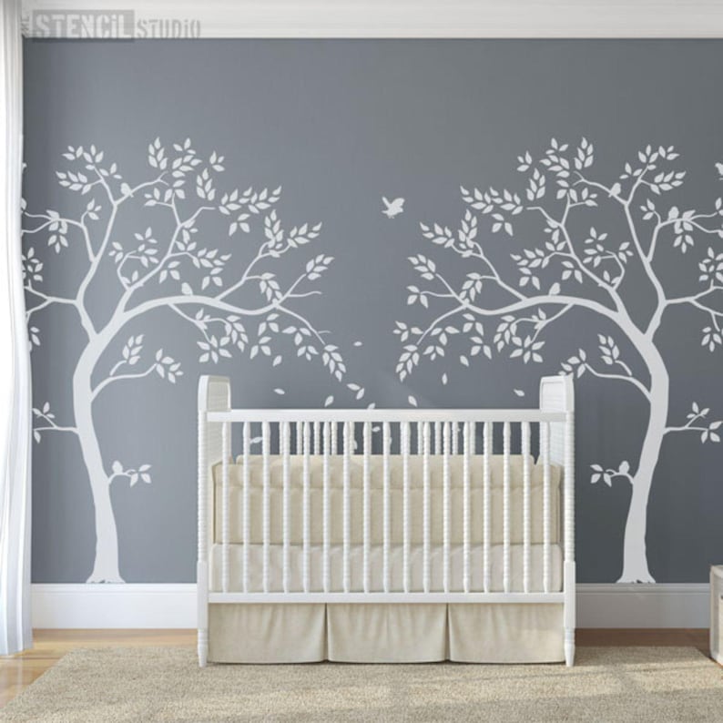 May include: A white crib with a beige mattress and skirt is centered in front of a gray wall. Two large white tree decals with leaves are on the wall. A bird decal is between the trees. The floor is covered with a beige rug.