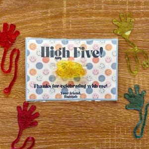May include: A wooden surface displays a card with "High Five!" text and smiley face pattern. Four colorful, hand-shaped toys in red, green, and yellow are arranged around the card. The card reads "Thanks for celebrating with me!" and "Your friend, Hannah."