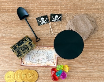 May include: A pirate-themed sensory kit featuring a black plastic shovel, two black skull and crossbones flags, a small treasure chest, a treasure map, a black container, a pile of brown kinetic sand, four gold coins, and six colorful plastic gems.