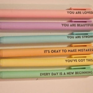 Puede incluir: Seis bolígrafos de colores pastel con mensajes inspiradores impresos en negro. Los bolígrafos son rosa, morado, azul, naranja, amarillo y verde. Los mensajes incluyen "You Are Loved", "You Are Beautiful" y "Every Day Is A New Beginning."