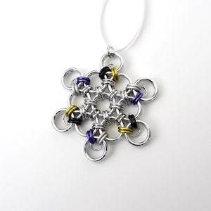 May include: A silver-colored snowflake pendant necklace with a white cord. The snowflake design is made of interlocking metal rings, with accents of black, yellow, and purple rings. The pendant is suspended from a thin, white cord.