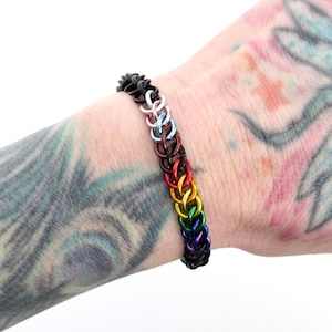 May include: A rainbow colored chainmail bracelet with black chainmail on either end. The bracelet is on a person's wrist.