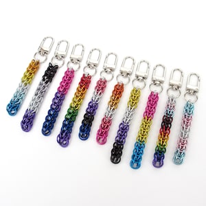 May include: A collection of ten chainmaille keychains, each featuring a silver-toned metal lobster clasp attached to a rectangular strip of interlocking jump rings. The rings are anodized in various vibrant color gradients, including blue, purple, pink, yellow, orange, and black, creating a distinct multi-colored pattern for each individual keychain.