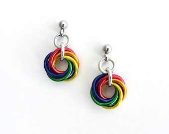 Könnte beinhalten: Ein Paar Ohrringe mit Regenbogenfarben. Jeder Ohrring hat einen kleinen, silberfarbenen Stecker, einen Verbindungsring und ein farbenfrohes, ineinander verschlungenes kreisförmiges Design. Die Farben umfassen Rot, Orange, Gelb, Grün und Blau.