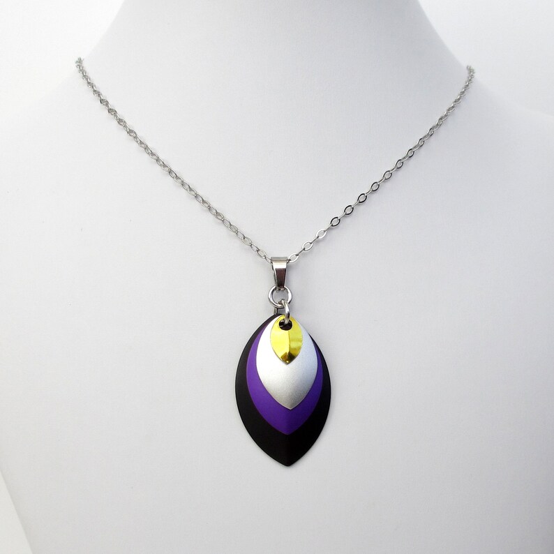 K&ouml;nnte beinhalten: Eine silberne Kettenkette mit einem Anh&auml;nger aus &uuml;bereinanderliegenden Metallbl&auml;ttern. Die Bl&auml;tter sind schwarz, lila, silber und gold.