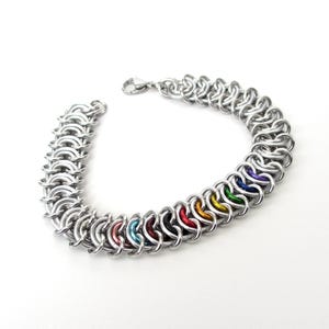 May include: A silver chainmail bracelet with a lobster clasp closure. The bracelet features a row of colorful rings in rainbow hues, including red, orange, yellow, green, blue, and purple. The bracelet is made of metal and is displayed on a white background.