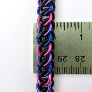 May include: Close-up of a black, blue, and pink chainmail bracelet measured against a ruler. The bracelet is approximately 2 3/4 inches long.