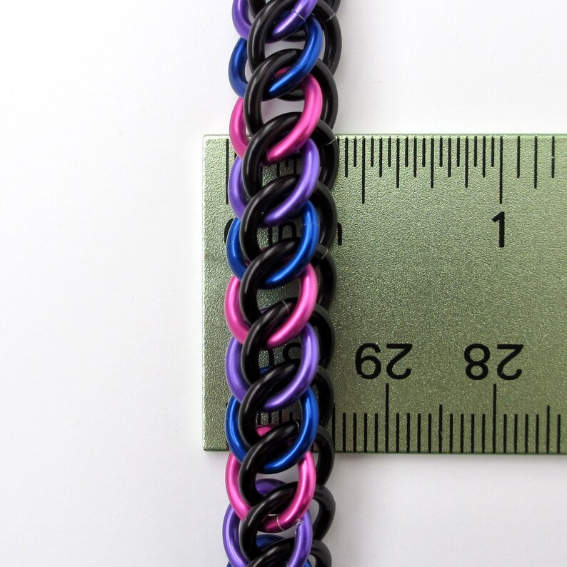 May include: Close-up of a black, blue, and pink chainmail bracelet measured against a ruler. The bracelet is approximately 2 3/4 inches long.