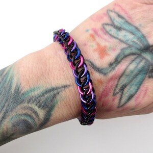 May include: A black, pink, and blue chainmail bracelet. The bracelet is made of metal rings that are linked together.