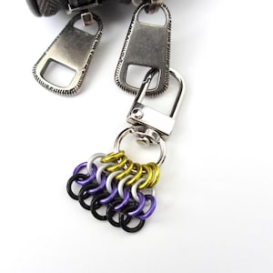 May include: A metal keychain charm featuring a chainmail weave pattern made of small interlocking rings in yellow, silver, purple, and black. The charm is attached to a silver-toned swivel lobster clasp, which is connected to two large zipper pulls.