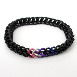 K&ouml;nnte beinhalten: Schwarzes Kettenhemdarmband mit gewebtem Design und einem Mittelteil aus f&uuml;nf Ringen in Pink, Wei&szlig;, Blau und Lila.