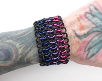 May include: A black, blue, pink, and purple chainmail bracelet. The bracelet is made of metal rings and is worn on a person's wrist.