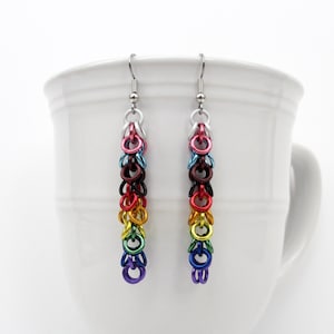May include: Rainbow colored dangle earrings with silver hooks. The earrings are made of small metal rings in a graduated size, with the largest rings at the bottom.