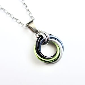 May include: A silver chain necklace with a pendant featuring three interlocking rings in black, gray, and green. The pendant is attached to a silver bail. The necklace is displayed against a white background.