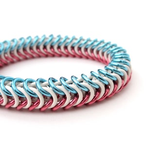 Peut inclure: Gros plan sur un bracelet en cotte de mailles. Le bracelet est composé d'anneaux métalliques entrelacés bleu clair, blancs et roses. Le bracelet est sur fond blanc.