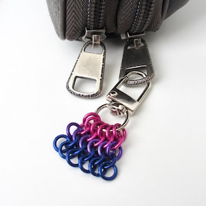 May include: A silver-toned metal keychain with a cluster of small, colorful metal rings in shades of blue, purple, and pink. The keychain is attached to a gray zippered pouch with silver hardware.