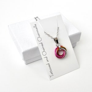 May include: A silver chain necklace with a pendant made of interlocking rings in pink, gold, and silver. The necklace is displayed on a white card with the text "Tattooed and Chained Chainmaille" and a white gift box.